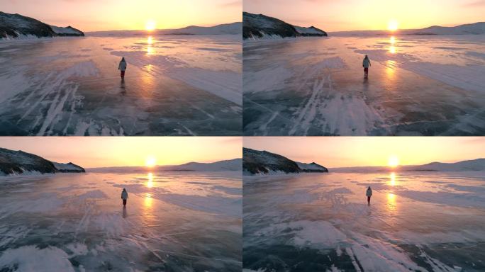 鸟瞰图上的年轻女子走在贝加尔湖的蓝色裂缝冰在美丽的橙色日出。太阳在冰面上的反射。贝加尔湖冰冻的冬季景