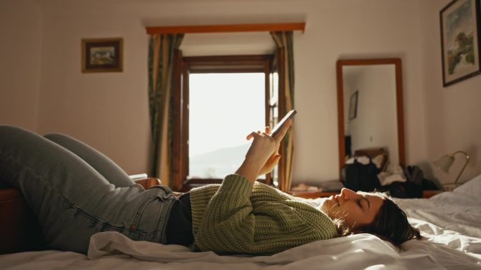 快乐的年轻女子在家里的床上通过智能手机自拍并在社交媒体应用上发布的侧视图