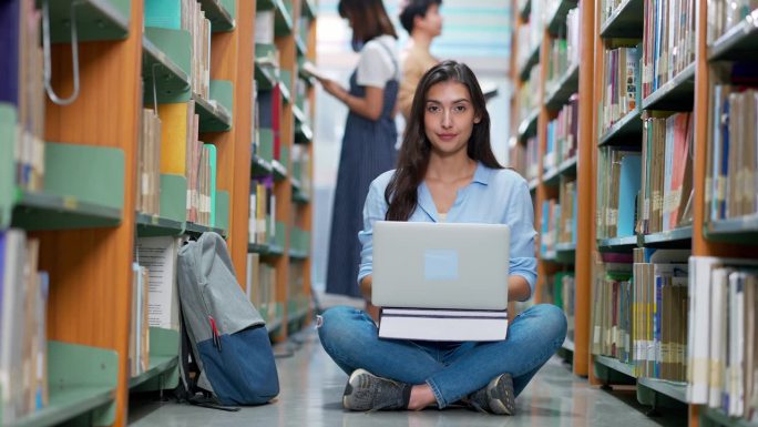 一名女学生坐在大学图书馆的书架过道里使用笔记本电脑
