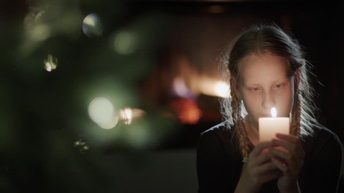 一个十几岁的女孩手里拿着一根燃烧的蜡烛。坐在背景的圣诞树旁，壁炉里正燃着火。在平安夜许个愿