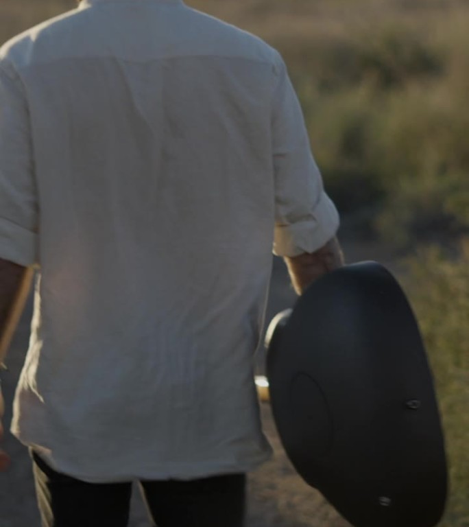 一个潮人拿着吉他走在夕阳下的草地上