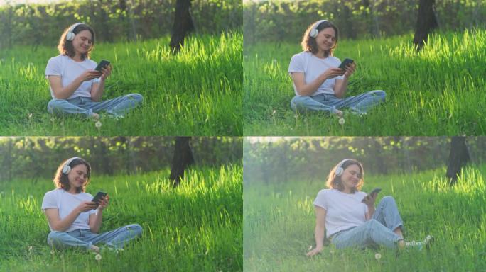 快乐、无忧无虑的女人戴着耳机坐在户外的绿草地上听音乐播客。年轻美丽的女孩在乡村享受美好的阳光。手持视