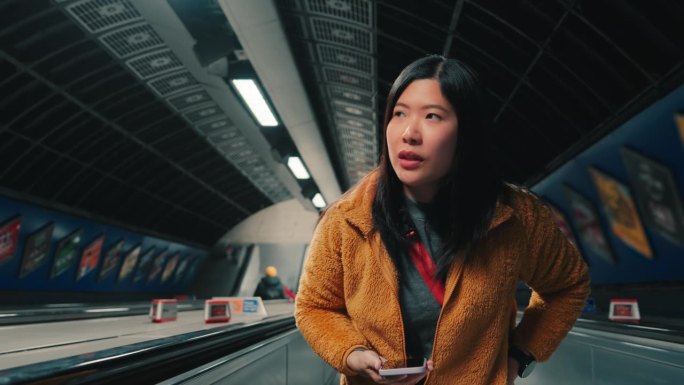 在英国伦敦地铁站的自动扶梯上，亚洲女游客一边用手机一边环顾四周的慢镜头