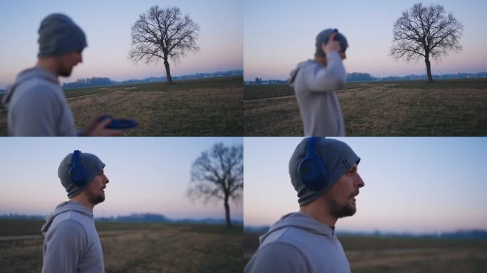 SLO - MO架对焦拍摄的单株光秃秃的树和戴着耳机的运动男子在蓝色小时热身前的田野
