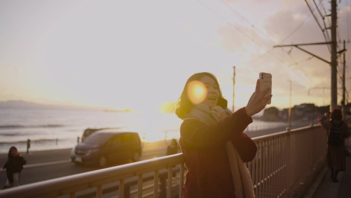 在夕阳的余晖中，一名年轻女子手持智能手机，在海岸边的铁路上进行视频通话时挥手，展示了令人惊叹的景色。
