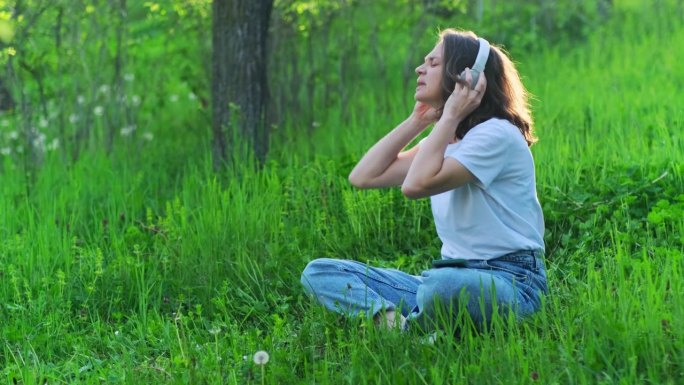 美丽的兴奋的女人戴着耳机，坐在户外阳光下的绿色草坪上听音乐，唱歌和跳舞。快乐的女性情绪。