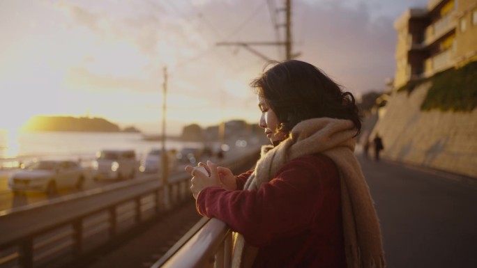 戴着围巾的年轻女子用手机拍摄日落。
