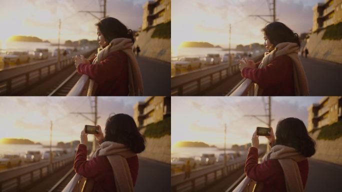 戴着围巾的年轻女子用手机拍摄日落。