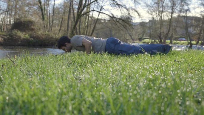 瘦骨嶙峋的年轻人在公园的河边绿草地上做俯卧撑。