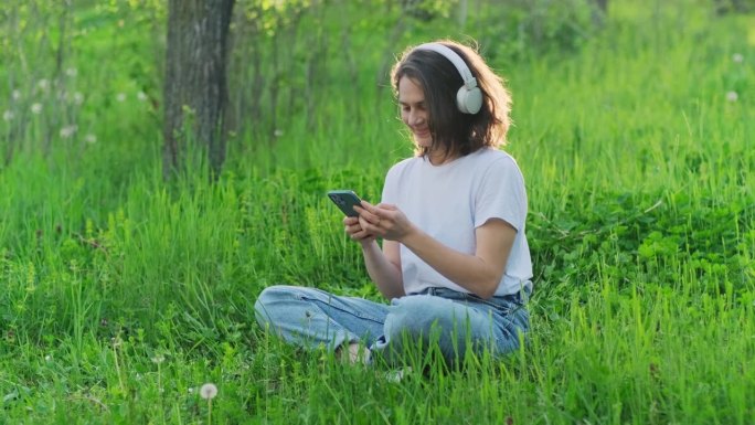 快乐、无忧无虑的女人戴着耳机坐在户外的绿草地上听音乐播客。年轻美丽的女孩在乡村享受美好的阳光。手持视