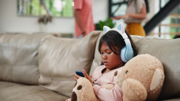 孩子、手机和耳机与父母在家里客厅的沙发上争吵、不和或离婚。在家庭冲突中，小女孩或女儿与泰迪熊玩手机