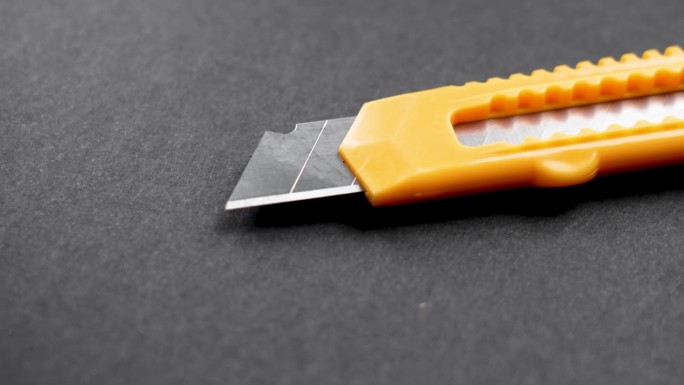 办公用刀，有钢制可伸缩的分段刀片，用于近距离切割黑色背景上的纸张。旋转