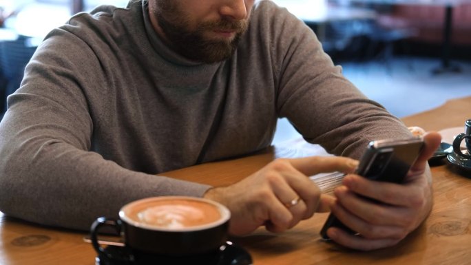 穿着灰色毛衣的成年男子坐在咖啡馆的木桌前玩智能手机。自由职业者的休息时间。时尚男士在舒适的咖啡馆氛围