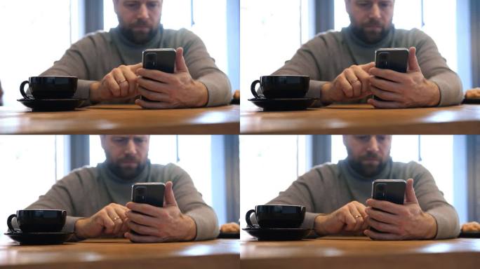 穿着灰色毛衣的成年男子坐在咖啡馆的木桌前玩智能手机。自由职业者的休息时间。时尚男士在舒适的咖啡馆氛围