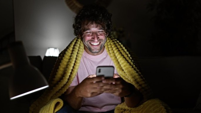 微笑的卷发男子晚上裹着黄毯子在室内使用智能手机