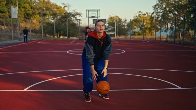 夏天的街头，一个穿着运动服和蓝色裤子的金发女孩，在地板上弹起一个橙色的篮球，用它做动作，然后把它扔进