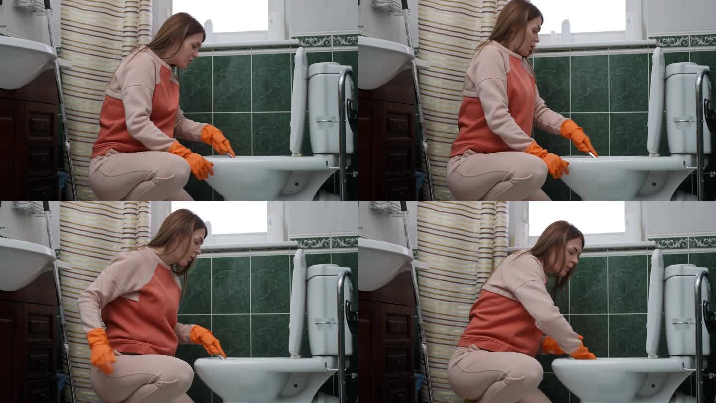 一位戴着橡胶手套的孕妇跪在现代住宅的浴室里，用马桶刷清洁马桶。女人累了，用袖子抹去额头上的汗。