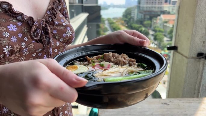 一个女孩手里拿着热汤，黑色的碗里盛着亚洲面汤、鸡肉、豆腐、蔬菜和鸡蛋的拉面。板岩的背景。前视图。