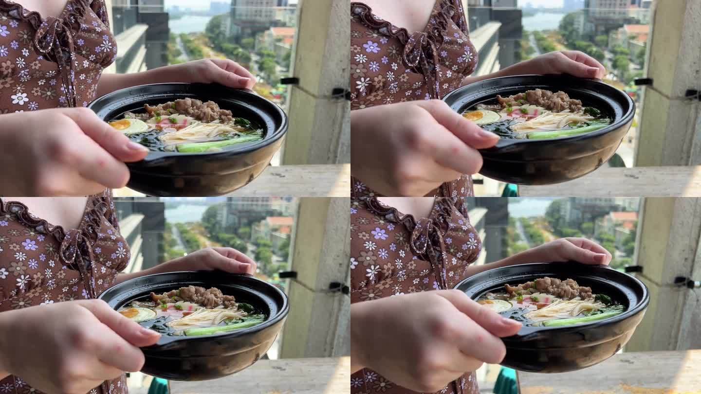 一个女孩手里拿着热汤，黑色的碗里盛着亚洲面汤、鸡肉、豆腐、蔬菜和鸡蛋的拉面。板岩的背景。前视图。