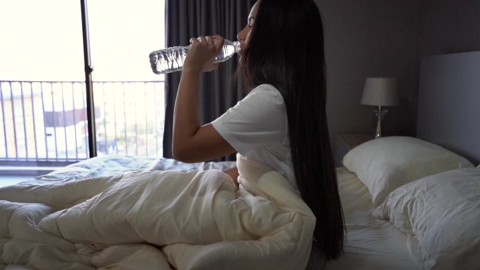 美丽的女人早上醒来，在卧室公寓的床上喝水。