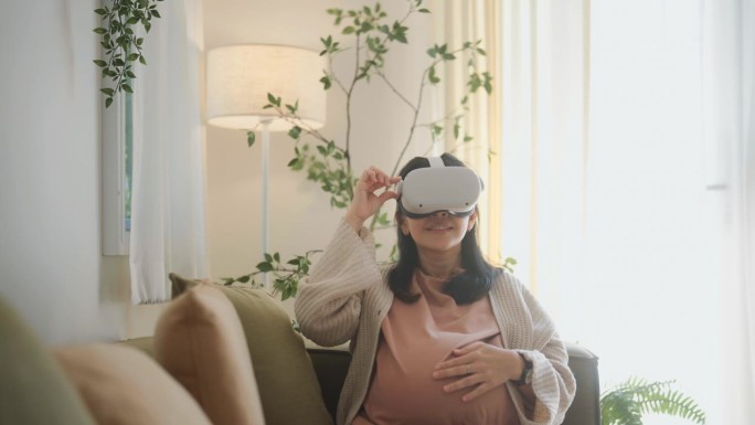 快乐的孕妇用虚拟现实眼镜练习教程照顾沙发上的新生婴儿。