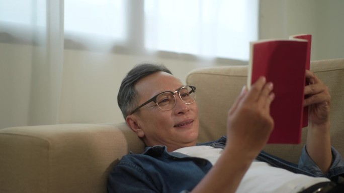 亚洲老年男子一边看书一边躺在家里的沙发上，安康退休男子躺在沙发上睡着，老年在家里休息舒适。