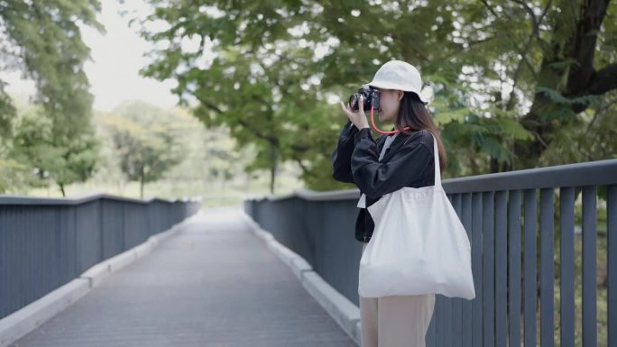年轻的亚洲女游客站在公园的桥上拿着相机拍照