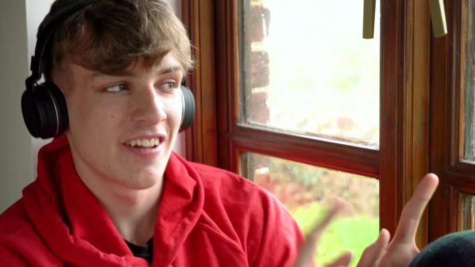 快乐微笑的男少年穿着红色连帽衫，坐在窗边用无线耳机和手机听音乐
