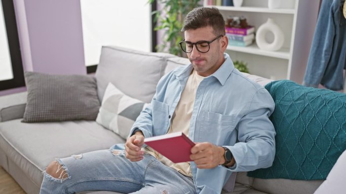 年轻的西班牙人坐在沙发上，戴着眼镜在家看书