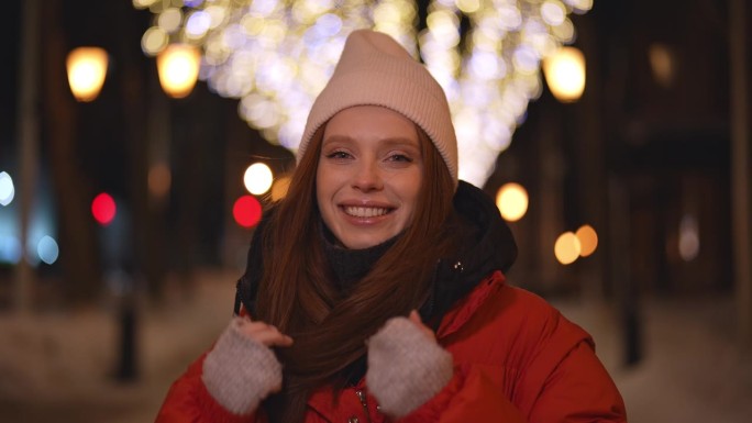 欢快的年轻女子戴着帽子，穿着红色的冬季夹克，站在雪城街道上，在铰链明亮的节日灯光照明的模糊背景上摆姿