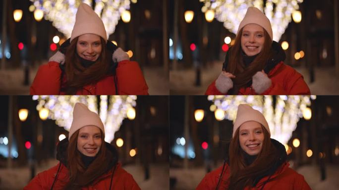 欢快的年轻女子戴着帽子，穿着红色的冬季夹克，站在雪城街道上，在铰链明亮的节日灯光照明的模糊背景上摆姿