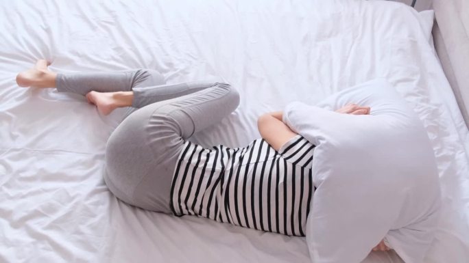 一个女人躺在床上因为疼痛，用枕头蒙住头。