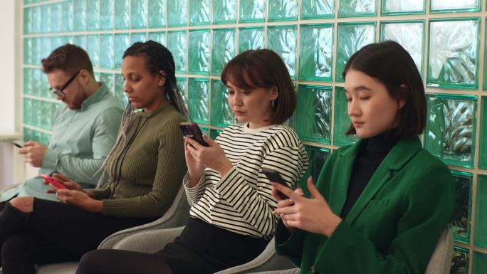 在现代化的办公大厅里，一群年轻的、不同种族的求职者正在使用智能手机等待面试。无聊的男性和女性空缺席位