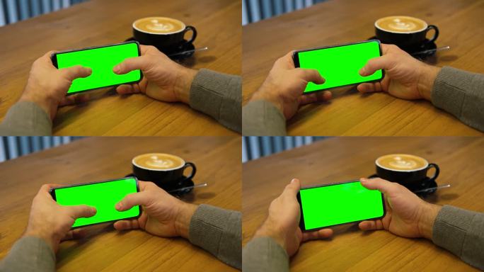 一个年轻人坐在咖啡馆里，手里拿着绿色的智能手机模型屏。男性使用色度键手机。水平模式。触摸，滑动显示屏