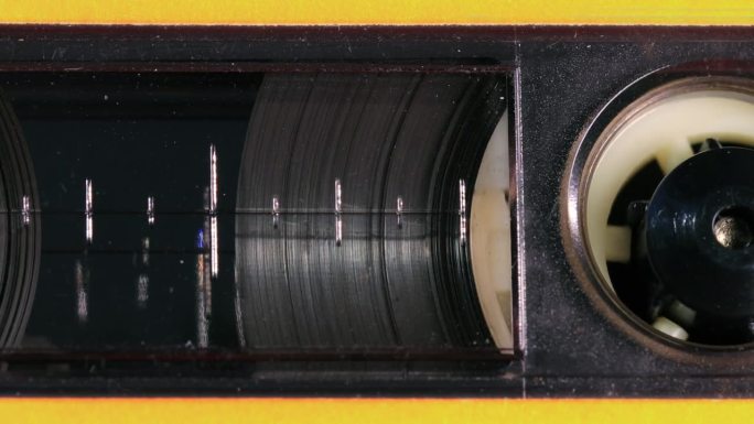 怀旧的盒式录音带在透明的窗户后面展开，带着黄色的塑料盖，磁带卷轴、百分比标记和其他细节随着镜头向左移