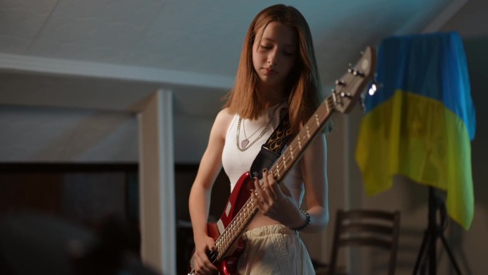 一名年轻的红发女孩站在一所音乐学校里，墙上挂着乌克兰国旗，她精力充沛地弹着贝斯吉他。