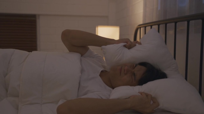 一个穿着睡衣的亚洲年轻人试图在漆黑的夜晚睡在床上。有魅力的男人躺在卧室里，因为噪音而感到心烦意乱和愤