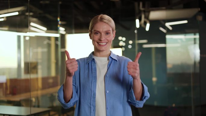 自信的白人女性竖起大拇指，对着玻璃墙背景上的镜头露出露齿的微笑。中年女商人做得很好，对潜在的同事表示