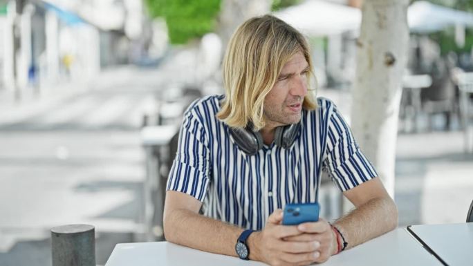 一位留着金色长发、戴着耳机的时尚成年男子坐在城市街道露台的桌子旁看他的智能手机。
