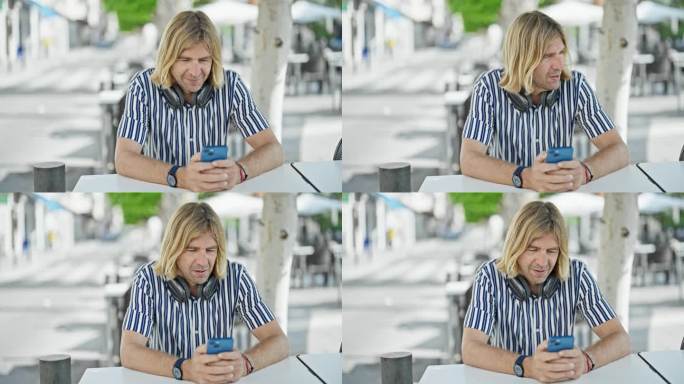 一位留着金色长发、戴着耳机的时尚成年男子坐在城市街道露台的桌子旁看他的智能手机。