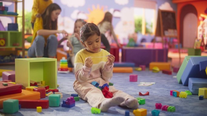 美丽的多民族小女孩坐在地板上玩五颜六色的积木玩具。一个可爱的亚洲孩子看着镜头微笑的肖像