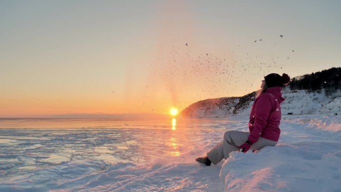 欢快的年轻女子在冰封的海面上抛起雪花，映衬着柔和的冬日夕阳。在美丽的冬季景色中玩耍