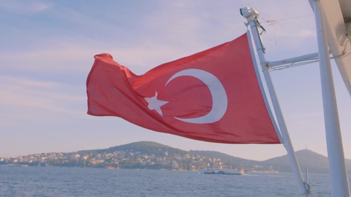 美丽的阳光下，土耳其国旗在风中飘扬的船巡航在夏天的大海和城市的背景