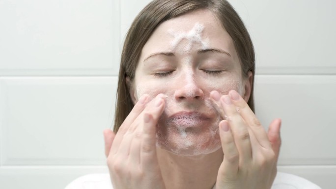 在白色瓷砖背景上，年轻女子将洁面泡沫涂抹在脸上。
