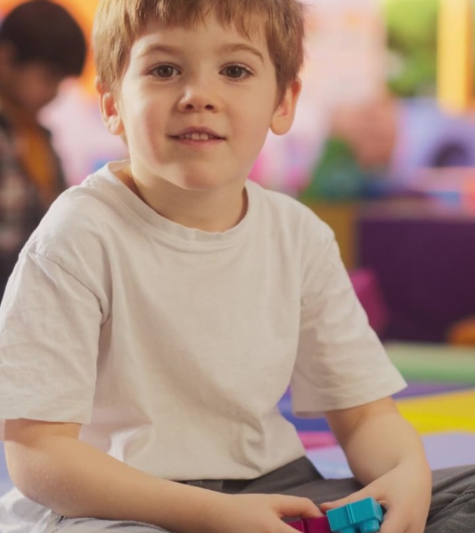 竖屏:一个好奇的白人男孩看着镜头微笑的肖像。在现代幼儿园，天真可爱的小男孩坐在地板上玩积木玩具