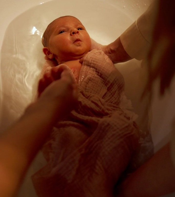 小婴儿躺在浴缸里的水里。妈妈的手在倒水。俯视图宝宝第一次洗澡。垂直视频