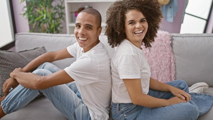 一对美丽的夫妇背靠背坐在舒适的沙发上，在他们可爱的家中快乐地微笑着，在他们牢固的关系中散发出积极的气