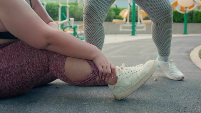 大码女性在公园朋友帮助和急救时腿部意外扭伤脚踝，胖女性在锻炼、活动和急救时脚部意外受伤。