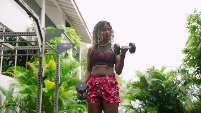 健美的混血女子在高档户外健身房里举哑铃，周围是茂密的热带绿色植物。运动女性增强肌肉张力，练习健身常规
