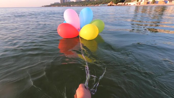 在一个阳光明媚的早晨，一个人在海岸线附近的海面上拉着一束用丝带系着的彩色小气球。慢动作